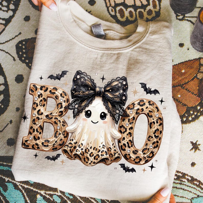 May include: A beige crewneck sweatshirt featuring a graphic print of the word BOO, where the letters B and O are filled with a brown leopard print pattern. A central ghost character wears a large black lace bow, surrounded by small black bats.