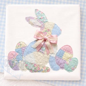 May include: A white square with a patchwork bunny and Easter eggs. The bunny is made of floral, gingham, and lace fabrics, with a pink bow. The eggs are pastel colors. The design is on a white background.
