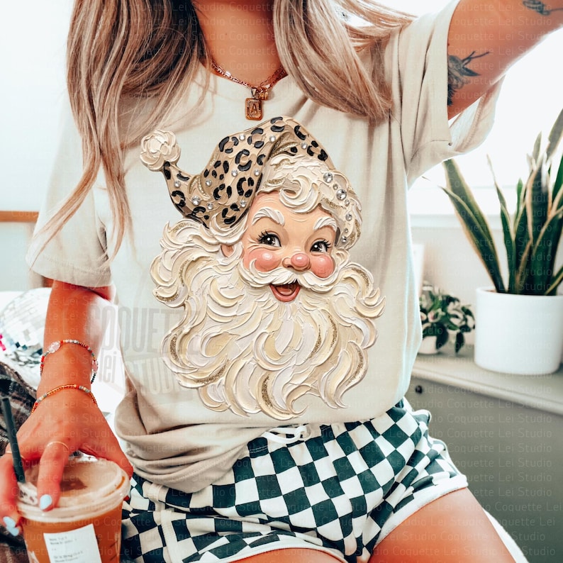 May include: A beige t-shirt featuring a graphic of a Santa Claus face with a thick white beard and a leopard print hat adorned with small rhinestones. The Santa illustration has a textured, painted appearance with rosy cheeks and a wide smile.