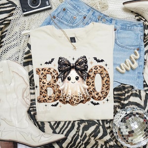 May include: A cream-colored t-shirt featuring a graphic of a ghost with a large black lace bow and leopard print accents. The text BOO is written in large leopard print letters. Small black bats and stars are scattered around the central design.