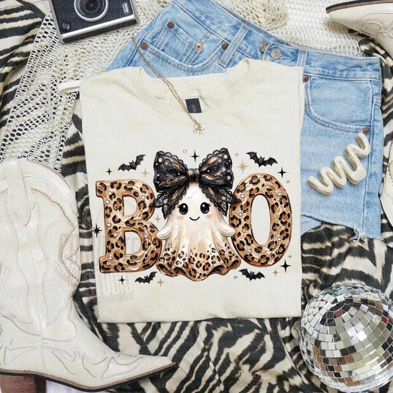 May include: A cream-colored t-shirt featuring a graphic of a ghost with a large black lace bow and leopard print accents. The text BOO is written in large leopard print letters. Small black bats and stars are scattered around the central design.