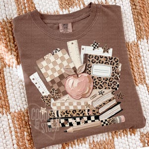 May include: Brown Comfort Colors crewneck sweatshirt with a school-themed graphic. The graphic includes a pink apple, books, notebooks, a pencil, and star accents. The design incorporates checkered and leopard print patterns.