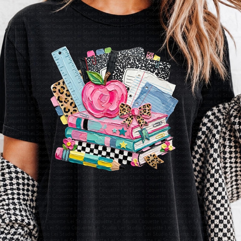 May include: Black t-shirt featuring a colorful school-themed graphic. The design includes a stack of books, a pink apple, a ruler, and a pencil. The books are decorated with stars and a leopard print bow.