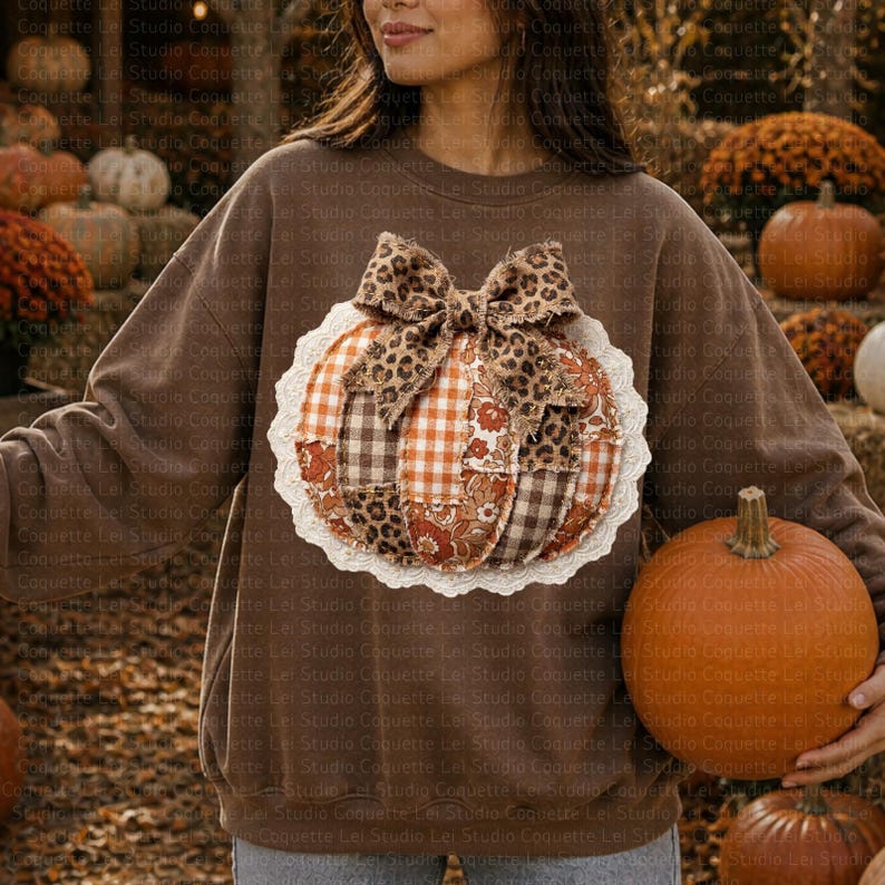 May include: Brown sweatshirt with a pumpkin applique. The pumpkin is made of various fabrics, including leopard print and plaid, and is trimmed with white lace. A large orange pumpkin is held in the hand.