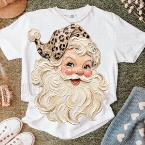 May include: A white short-sleeve t-shirt featuring a large graphic of a Santa Claus face with a thick, textured white beard and a leopard print hat adorned with small clear rhinestones. The Santa illustration has a cheerful expression.
