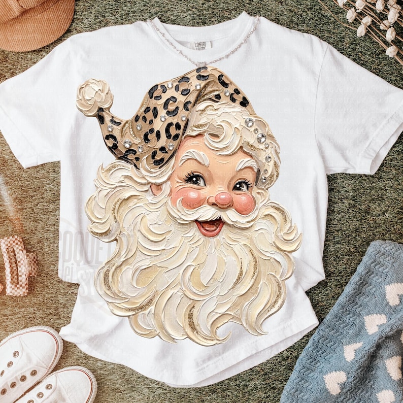 May include: A white short-sleeve t-shirt featuring a large graphic of a Santa Claus face with a thick, textured white beard and a leopard print hat adorned with small clear rhinestones. The Santa illustration has a cheerful expression.