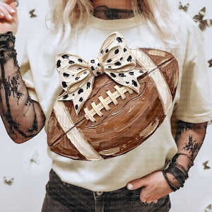 May include: A cream-colored t-shirt featuring a graphic of a brown American football with a white and black dalmatian-spotted bow on top. The graphic has a textured, painted appearance with visible brushstrokes and white laces on the football.