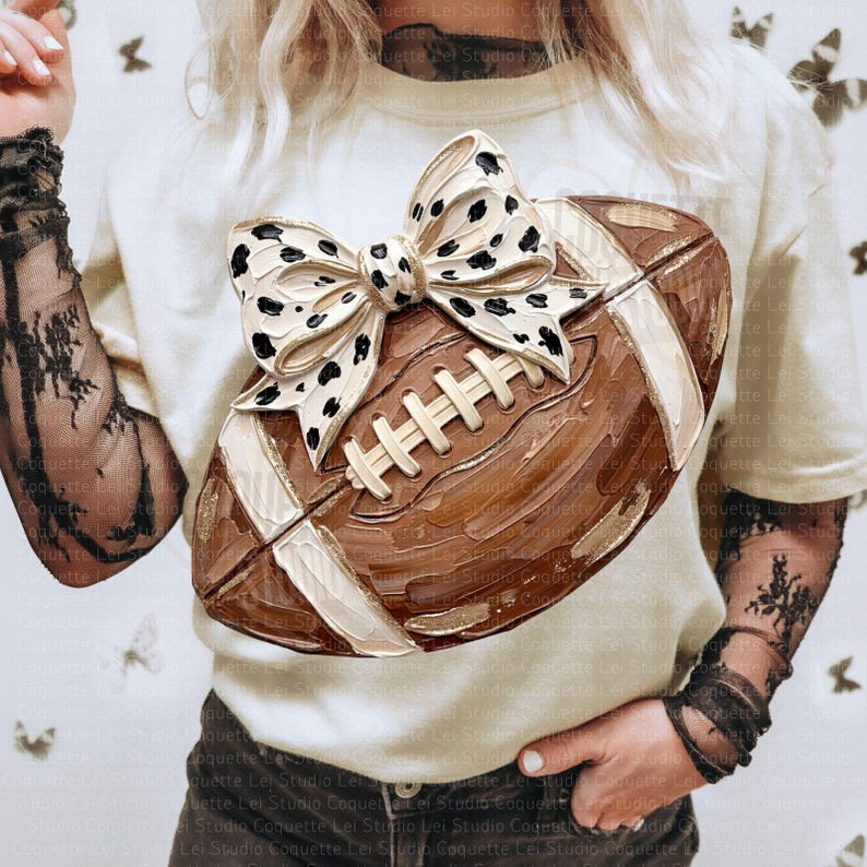 May include: A cream-colored t-shirt featuring a graphic of a brown American football with a white and black dalmatian-spotted bow on top. The graphic has a textured, painted appearance with visible brushstrokes and white laces on the football.