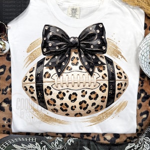 May include: A white t-shirt featuring a graphic of a football with a tan and black leopard print pattern, accented by a large black bow adorned with clear rhinestones. The design is framed by gold glitter brushstrokes on the fabric.