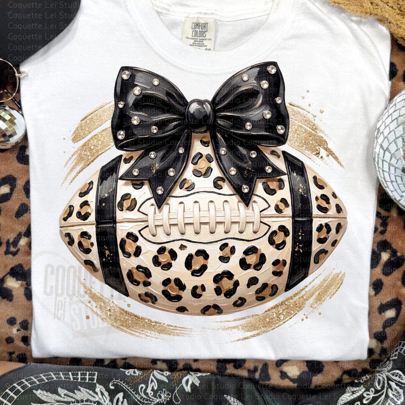 May include: A white t-shirt featuring a graphic of a football with a tan and black leopard print pattern, accented by a large black bow adorned with clear rhinestones. The design is framed by gold glitter brushstrokes on the fabric.