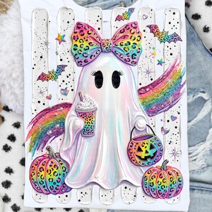 May include: A white t-shirt featuring a central graphic of a ghost with a rainbow leopard print bow, holding a matching leopard print cup and a jack-o'-lantern bucket. The design includes rainbow streaks, bats, stars, and two leopard print pumpkins.