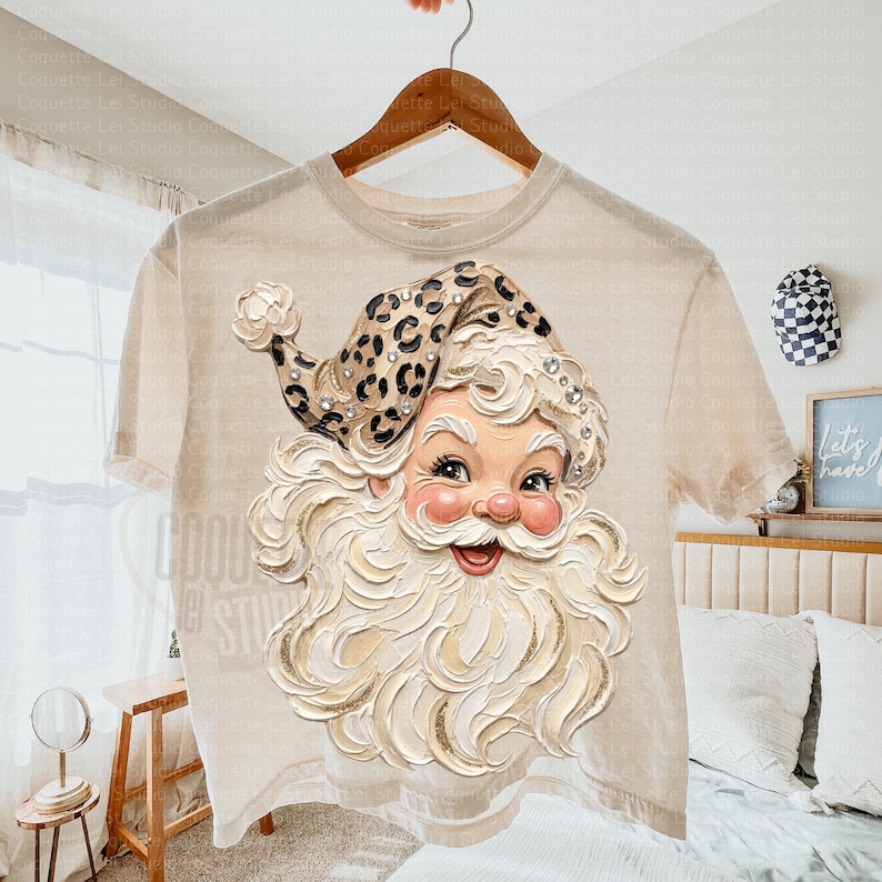 May include: A beige short-sleeve t-shirt featuring a graphic of a Santa Claus face with a thick white beard and a leopard print hat. The Santa illustration includes small sparkling accents throughout the hat and beard, set against a neutral fabric.