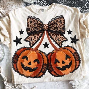 May include: Cream-colored t-shirt featuring a Halloween design. The design includes two orange jack-o'-lanterns with black lace trim, a leopard print bow, and black star accents. The shirt has long sleeves.