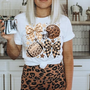 May include: A white short-sleeve t-shirt featuring a graphic print of a football, a football helmet, and two bows, all decorated with leopard print and checkered patterns. The shirt is paired with brown and black leopard print leggings.