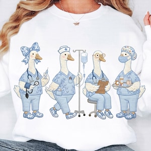 Puede incluir: Sudadera blanca con un diseño caprichoso de cuatro gansos de dibujos animados vestidos como profesionales médicos. Cada ganso lleva uniformes azules claros y accesorios como estetoscopios, jeringas y mascarillas. Los gansos están en varias poses.
