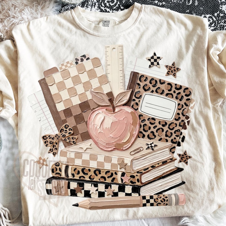 May include: Cream-colored t-shirt featuring a stack of books, an apple, and school supplies. The design includes a pencil, ruler, and notebook with a leopard print and checkered patterns. The shirt has a Comfort Colors label.