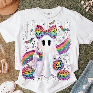 May include: A white t-shirt featuring a central graphic of a ghost with a large, colorful leopard-print bow, holding a patterned cup and a jack-o'-lantern bucket. The design includes rainbow wings, colorful pumpkins, and bats on a striped background.