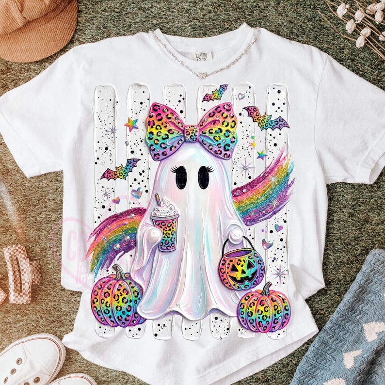 May include: A white t-shirt featuring a central graphic of a ghost with a large, colorful leopard-print bow, holding a patterned cup and a jack-o'-lantern bucket. The design includes rainbow wings, colorful pumpkins, and bats on a striped background.