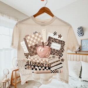 May include: Beige t-shirt with a school-themed graphic. The design features a stack of books, a pink apple, a ruler, and a pencil. The books have checkered and leopard print patterns. The shirt is hanging on a wooden hanger.