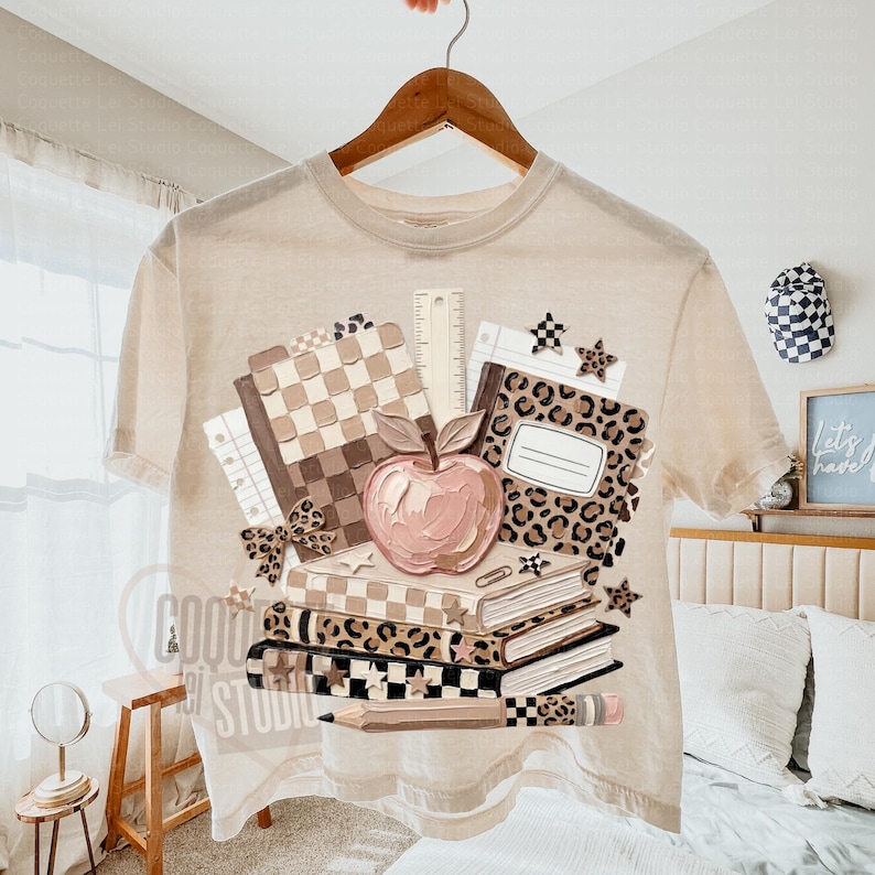May include: Beige t-shirt with a school-themed graphic. The design features a stack of books, a pink apple, a ruler, and a pencil. The books have checkered and leopard print patterns. The shirt is hanging on a wooden hanger.