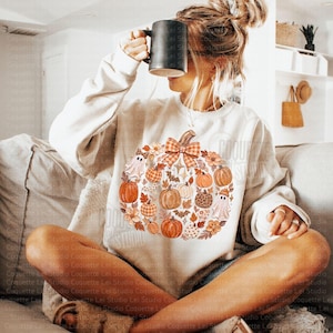 May include: A cream-colored crewneck sweatshirt featuring a large central graphic of a pumpkin composed of smaller pumpkins, ghosts, flowers, leaves, bats, and a plaid bow. The sweatshirt has long sleeves and a ribbed neckline and cuffs.