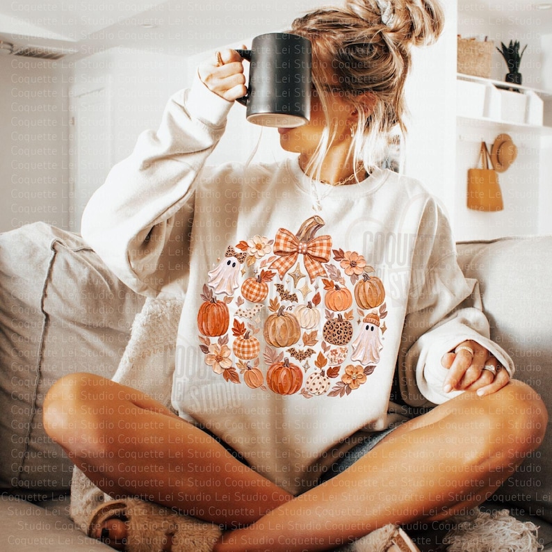 May include: A cream-colored crewneck sweatshirt featuring a large central graphic of a pumpkin composed of smaller pumpkins, ghosts, flowers, leaves, bats, and a plaid bow. The sweatshirt has long sleeves and a ribbed neckline and cuffs.