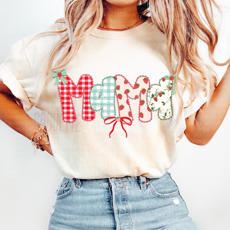 May include: Cream-colored t-shirt featuring the word "MAMA" in a patchwork design. The letters are in red gingham, light blue plaid, pink with strawberries, and floral patterns. The design includes bows and a red ribbon.