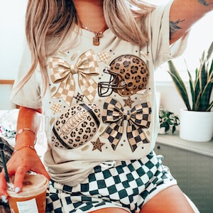 May include: A cream-colored t-shirt featuring a graphic print of two large checkered bows, a leopard-print football helmet, a leopard-print football, and several small stars. The design is centered on the front of the shirt.