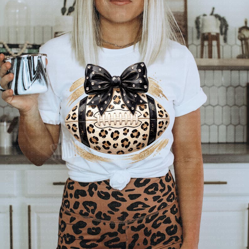 May include: A white short-sleeve t-shirt featuring a graphic of an American football with a leopard print pattern, topped with a large black bow with white polka dots. The design is centered on the chest and includes gold brushstroke accents.