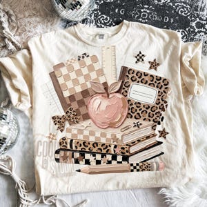May include: Cream-colored t-shirt featuring a school-themed graphic. The design includes a stack of books, an apple, a pencil, and notebooks with checkered and leopard print patterns. The text "COOL lei STUDIO" is visible.