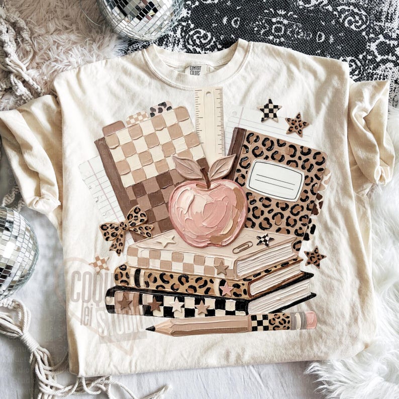 May include: Cream-colored t-shirt featuring a school-themed graphic. The design includes a stack of books, an apple, a pencil, and notebooks with checkered and leopard print patterns. The text "COOL lei STUDIO" is visible.