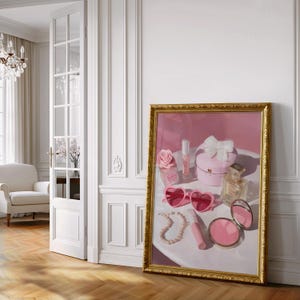 May include: A framed artwork featuring a still life of pink accessories. The painting includes heart-shaped sunglasses, a pearl necklace, a perfume bottle, a compact mirror, and a gift box with a bow. The frame is gold.