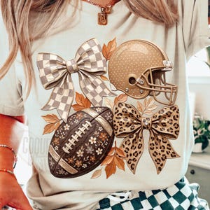 Peut inclure: Un t-shirt beige avec une impression graphique représentant un casque de football américain à pois blancs, un ballon de football à motifs floraux blancs, un nœud à carreaux, un nœud léopard et des feuilles d'automne orange dispersées.