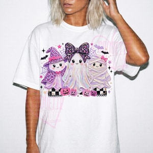 May include: A white t-shirt featuring a graphic of three cartoon ghosts in pastel colors. One ghost wears a leopard print witch hat, the center ghost has a large leopard print bow, and the third is wrapped like a mummy. Small pink pumpkins are below.