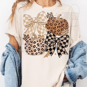May include: A cream-colored t-shirt featuring a graphic print of two pumpkins with leopard print patterns, two large bows with checkered patterns in beige and black, and small autumn leaves, layered under a blue denim jacket.