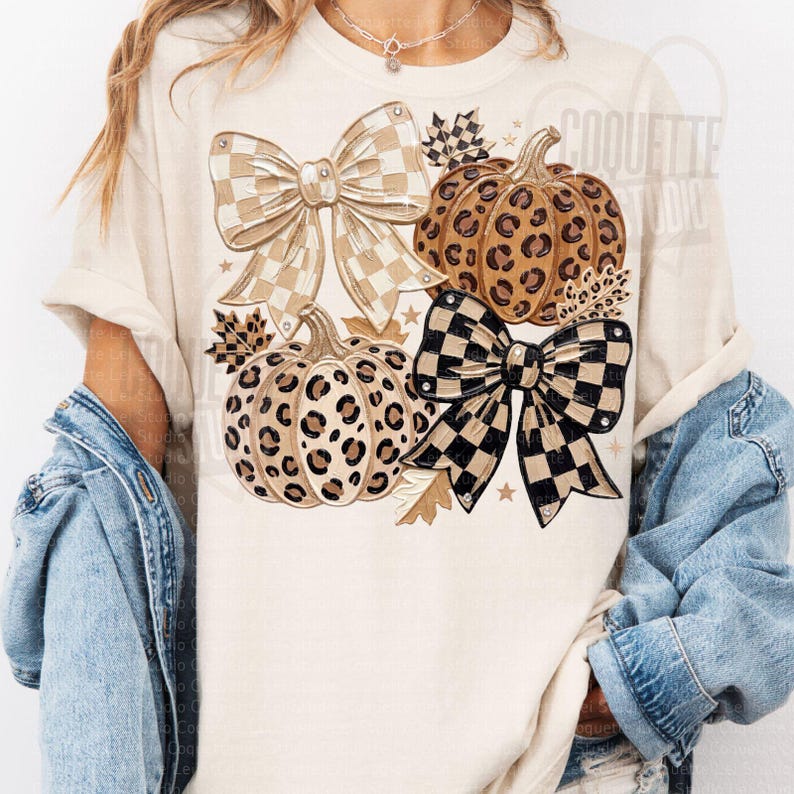 May include: A cream-colored t-shirt featuring a graphic print of two pumpkins with leopard print patterns, two large bows with checkered patterns in beige and black, and small autumn leaves, layered under a blue denim jacket.