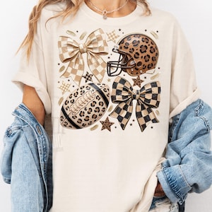May include: A cream-colored t-shirt featuring a graphic print of two checkered bows, a leopard print football helmet, a leopard print football, and several small stars. The design uses a neutral color palette of tan, brown, and black throughout.