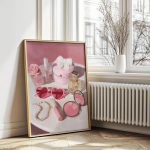 May include: A framed artwork featuring a still life of pink accessories. The composition includes heart-shaped sunglasses, a pearl necklace, a compact mirror, perfume bottles, and a gift box with a bow. A rose-shaped perfume bottle and lipstick are also visible.