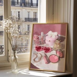 May include: A framed print featuring a collection of pink accessories. The image includes heart-shaped sunglasses, a pearl necklace, perfume bottles, a compact, and a box with a bow. A vase of white flowers sits in front of the print.
