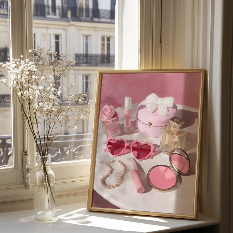 May include: A framed print featuring a collection of pink accessories. The image includes heart-shaped sunglasses, a pearl necklace, perfume bottles, a compact, and a box with a bow. A vase of white flowers sits in front of the print.