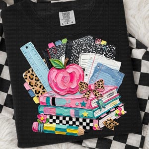 May include: Black crew neck sweatshirt with a colorful school-themed design. The design features a stack of books, an apple, a ruler, and notebooks. The design includes pink, blue, yellow, and leopard print accents. The sweatshirt has a Comfort Colors tag.