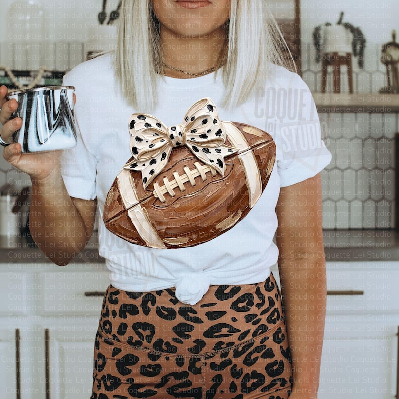 May include: A white short-sleeve t-shirt featuring a brown football graphic adorned with a large leopard print bow. The shirt is paired with brown and black leopard print bottoms. A white and black marbled mug is held in the foreground.