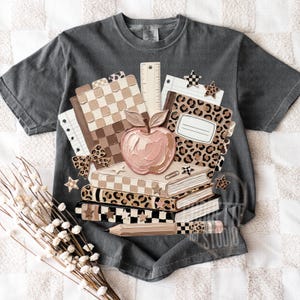 May include: Dark gray t-shirt featuring a school-themed graphic. The design includes a stack of books, a pink apple, a ruler, and notebooks with checkered and leopard print patterns. The graphic also has star and bow accents.