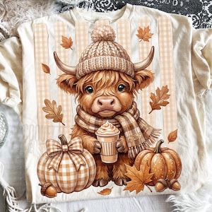 Può includere: Una t-shirt color crema con la grafica di una soffice mucca delle Highland che indossa un berretto di lana e una sciarpa a quadri, mentre tiene in mano una tazza di caffè. Il design include due zucche, una a quadri con fiocco, e foglie autunnali.