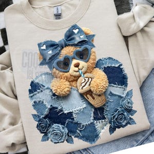 May include: Beige sweatshirt featuring a teddy bear wearing heart-shaped sunglasses and a denim bow. The bear is holding a drink and is set against a denim heart with denim roses. The text "COO" is visible.