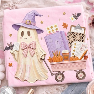 May include: A light pink t-shirt featuring a graphic of a cream-colored ghost wearing a purple witch hat and a pink plaid bow. The ghost pulls a pink wagon filled with a purple notebook, pencils, pumpkins, a coffee cup, and graded papers.