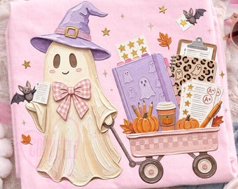 May include: A light pink t-shirt featuring a graphic of a cream-colored ghost wearing a purple witch hat and a pink plaid bow. The ghost pulls a pink wagon filled with a purple notebook, pencils, pumpkins, a coffee cup, and graded papers.