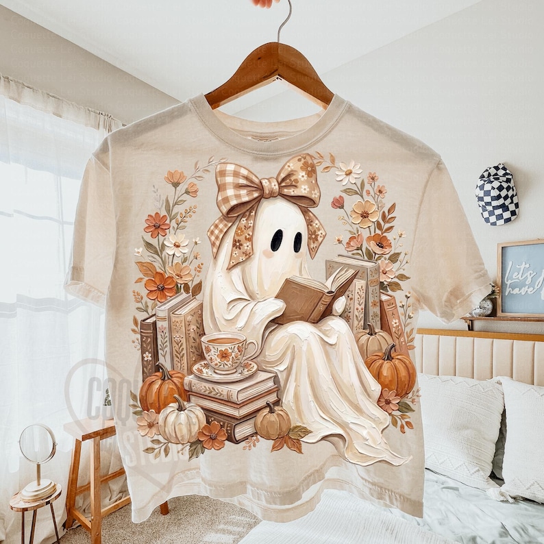 Puede incluir: Una camiseta beige de manga corta con un estampado gr&aacute;fico de un fantasma blanco con un lazo a cuadros marr&oacute;n, leyendo un libro entre pilas de libros, calabazas y arreglos florales en tonos naranja, marr&oacute;n y crema.