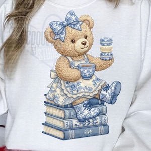 May include: A white sweatshirt with a teddy bear design. The bear wears a blue floral bow, apron, and boots, holding a teacup and macarons. The bear sits on a stack of books with blue floral patterns.