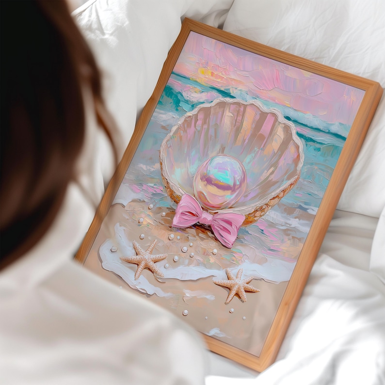 Puede incluir: Una obra de arte enmarcada que presenta una concha con una gran perla iridiscente y un lazo rosa. La pintura representa una escena de playa con estrellas de mar y perlas en la arena, con un cielo pastel y un oc&eacute;ano de fondo. El marco es de madera clara.